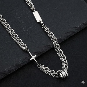 May include: A silver-toned necklace with multiple chain layers. The necklace features a cross pendant with a small gem and a rectangular tag. The design includes a series of interconnected links and a decorative cylindrical element.