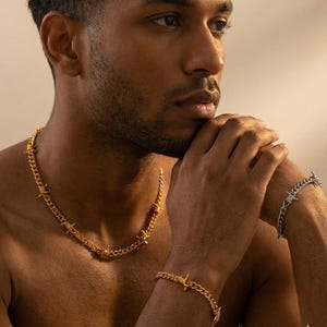 May include: A gold-coloured barbed wire necklace and bracelet set. A silver-coloured barbed wire bracelet is also visible. The jewellery is worn on a person's neck and wrist. The jewellery is made of metal.