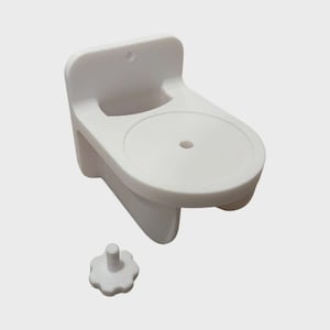 May include: A white plastic wall-mounted soap dish with a round soap holder and a small, separate, white, flower-shaped knob. The dish has a back and two side supports.