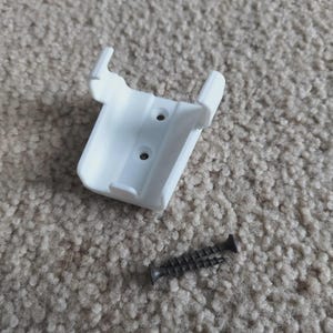 May include: A white plastic bracket with two screw holes and a black screw. The bracket has two curved arms and two small tabs. The screw is dark grey and has a pointed end.