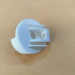 Peut inclure: Un objet blanc imprimé en 3D, avec une base cylindrique et une section supérieure rectangulaire. La section supérieure comporte un trou central et deux barres horizontales. L'objet semble être un composant ou une pièce d'un mécanisme plus grand.