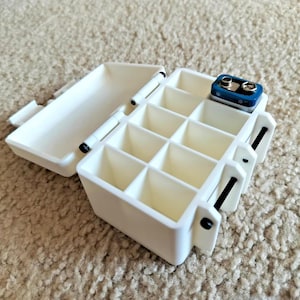 Puede incluir: Un organizador de baterías de plástico blanco, abierto para revelar una cuadrícula de compartimentos. Una batería de 9 voltios azul y plateada está en un compartimento. La caja tiene una tapa con bisagras y bisagras negras.