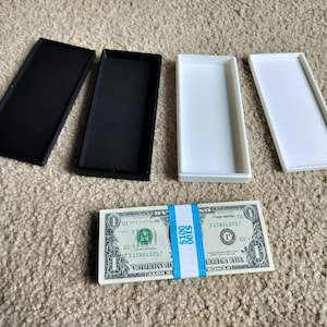 Op de afbeelding: Een bundel van één dollar biljetten, vastgezet met een blauw-witte papieren band, naast twee zwarte en twee witte rechthoekige trays. De band leest "$100" en "$100 DOLLARS".