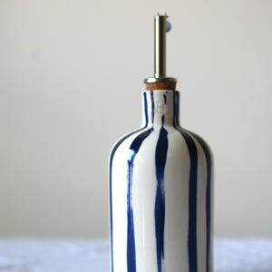 Könnte beinhalten: Eine weiße Keramikflasche mit vertikalen blauen Streifen, gekrönt von einem Korken und einem Edelstahl-Ausgießer. Die Flasche ist zum Ausgießen von Flüssigkeiten wie Öl oder Essig bestimmt.