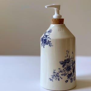 Könnte beinhalten: Ein weißer Keramik-Seifenspender mit blauem Blumenmuster. Der Spender hat eine weiße Pumpe und einen Korken. Das Design erinnert an Vintage-Porzellan. Geeignet für Bad oder Küche.