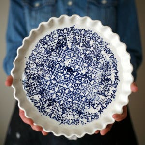 Puede incluir: Fuente de tarta de cerámica blanca con un estampado floral azul. La fuente tiene un borde festoneado y está sostenida por una persona. El patrón azul cubre el interior.