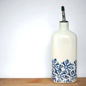 Może przedstawiać: Biała ceramiczna butelka na olej ze stalowym dozownikiem. Dół butelki zdobi niebieski kwiatowy wzór. Butelka jest prostokątna z zaokrąglonymi krawędziami i stoi na drewnianej powierzchni.