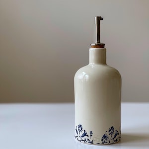 May include: Cream-coloured ceramic oil bottle with a stainless steel spout and cork stopper. The bottle has a blue floral design around the base. A decorative and functional kitchenware item.