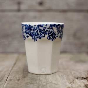 May include: White ceramic tumbler with a faceted design. The top edge features a detailed blue floral pattern. The cup sits on a wooden surface, showcasing a simple, elegant design. A small square mark is visible on the bottom.