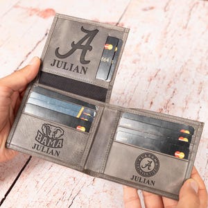 May include: Gray leather wallet with multiple card slots. The wallet is open, revealing several credit cards. The wallet is personalized with the name "JULIAN" and the letter "A" and the words "BAMA" and "ALABAMA CRIMSON TIDE".