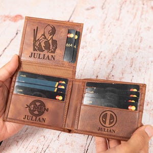 May include: Open brown leather wallet with multiple card slots. The wallet features engraved designs including a Deadpool logo, X-Men logo, and a character image. The name "JULIAN" is also engraved on the wallet.