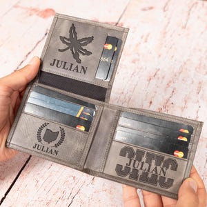 May include: Gray leather wallet with multiple card slots, open to display contents. The wallet is personalized with the name "JULIAN" and various designs, including a leaf, a state outline, and a stylized text. Several credit cards are visible inside.