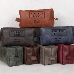 May include: A collection of personalized leather toiletry bags in various colors, including brown, gray, and blue. Each bag features a rectangular label with the text "COLLEGE LOGO", the name "FRANK", and "Class of 2026".