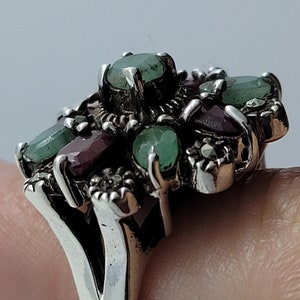 May include: A silver ring with a cluster of green and red gemstones. The ring is set with emerald and ruby stones, and has a delicate floral design.