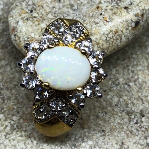 Puede incluir: Un anillo de oro con una piedra de ópalo blanco rodeada de piedras preciosas más pequeñas y transparentes.
