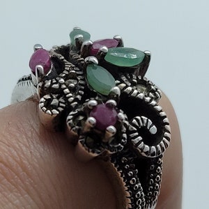 Può includere: Un anello d'argento con un gruppo di pietre preziose verdi e rosse incastonate in un design filigranato dettagliato.