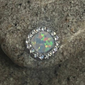 Peut inclure: Une bague en argent avec une pierre d'opale ronde entourée de petites pierres claires.