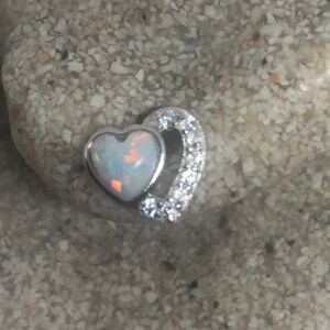 May include: A silver pendant featuring two interlocking hearts. The larger heart is set with a white opal gemstone, while the smaller heart is accented with clear cubic zirconia stones.