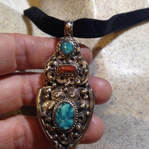 May include: A silver pendant necklace with a black velvet choker. The pendant features intricate filigree work and three turquoise and coral stones.