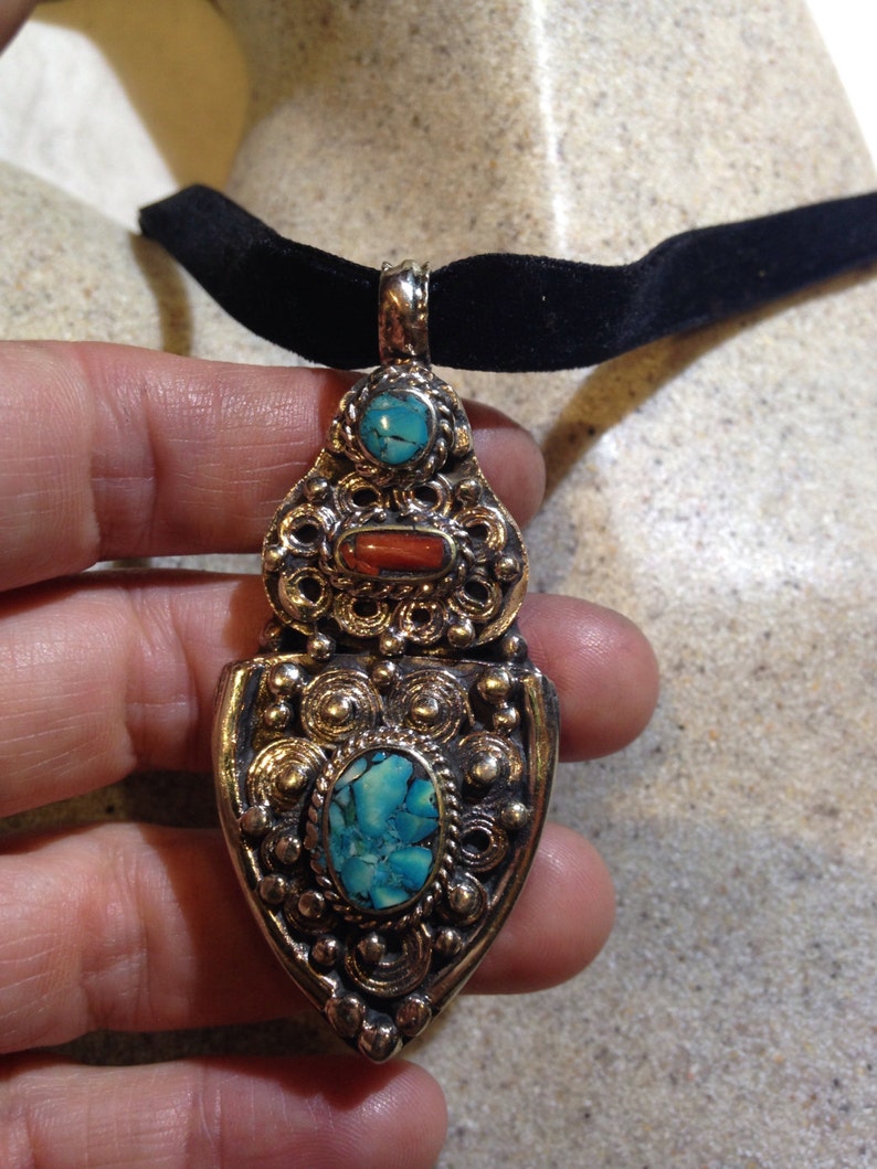 May include: A silver pendant necklace with a black velvet choker. The pendant features intricate filigree work and three turquoise and coral stones.