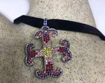 May include: A silver-toned cross pendant necklace with a black velvet choker. The cross is adorned with red, blue, and yellow gemstones. The pendant is a decorative piece of jewelry.