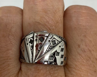 Puede incluir: Un anillo de plata con un diseño de cartas de juego, incluyendo el As, el Rey, la Reina, el Jack y el 10 de diamantes. El anillo se lleva en el dedo de una persona.