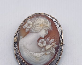 May include: A cameo brooch featuring a woman's profile with a flower in her hair, set in a gold filigree frame.