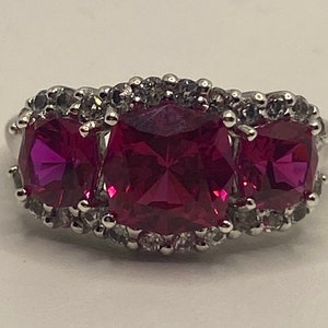 Pode incluir: Um anel de prata com três pedras preciosas grandes de forma quadrada de cor rosa. Cada pedra é rodeada por um halo de pedras preciosas menores e transparentes.