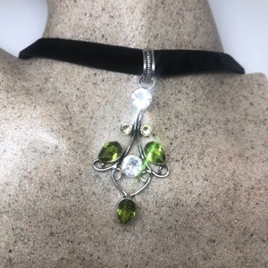 May include: A black velvet choker necklace with a silver pendant featuring clear and green gemstones. The pendant is in the shape of a stylized flower with a teardrop-shaped gemstone hanging from the bottom.