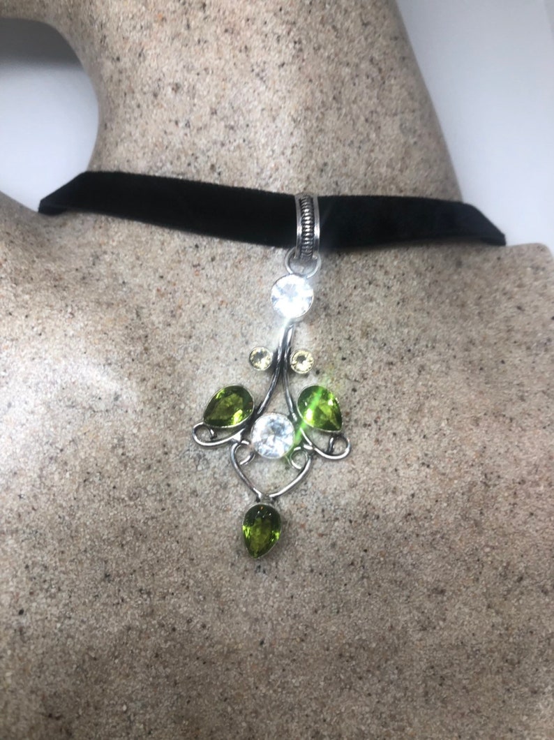 May include: A black velvet choker necklace with a silver pendant featuring clear and green gemstones. The pendant is in the shape of a stylized flower with a teardrop-shaped gemstone hanging from the bottom.
