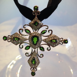 Puede incluir: Un colgante de cruz de plata con piedras preciosas verdes y transparentes. La cruz es adornada con detalles intrincados y está suspendida de una cinta negra.