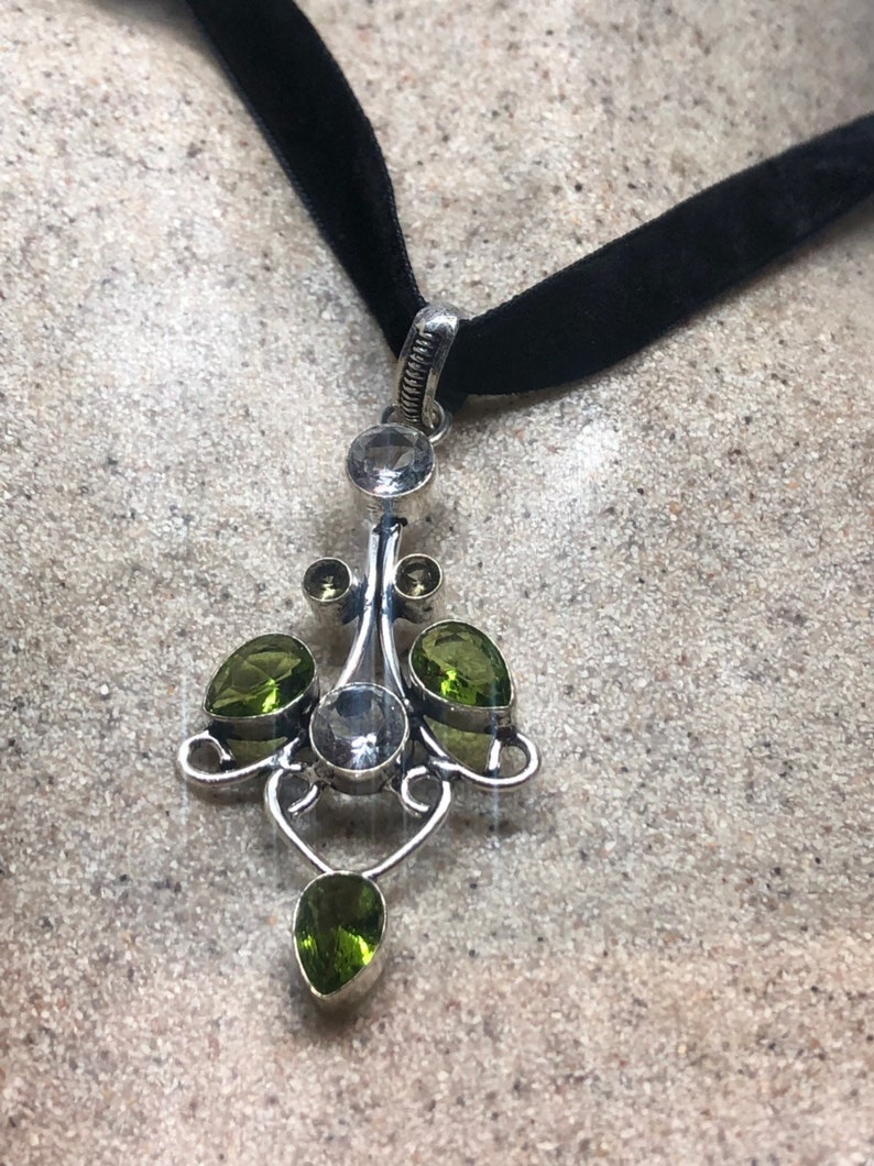 May include: A silver pendant necklace with a black velvet choker. The pendant features a delicate design with clear and green gemstones.