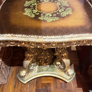 May include: Ornate, square-topped table with a gold-colored, decorative base. The tabletop is brown with a gold-colored floral design in the center. The table is likely made of wood and is a piece of furniture.