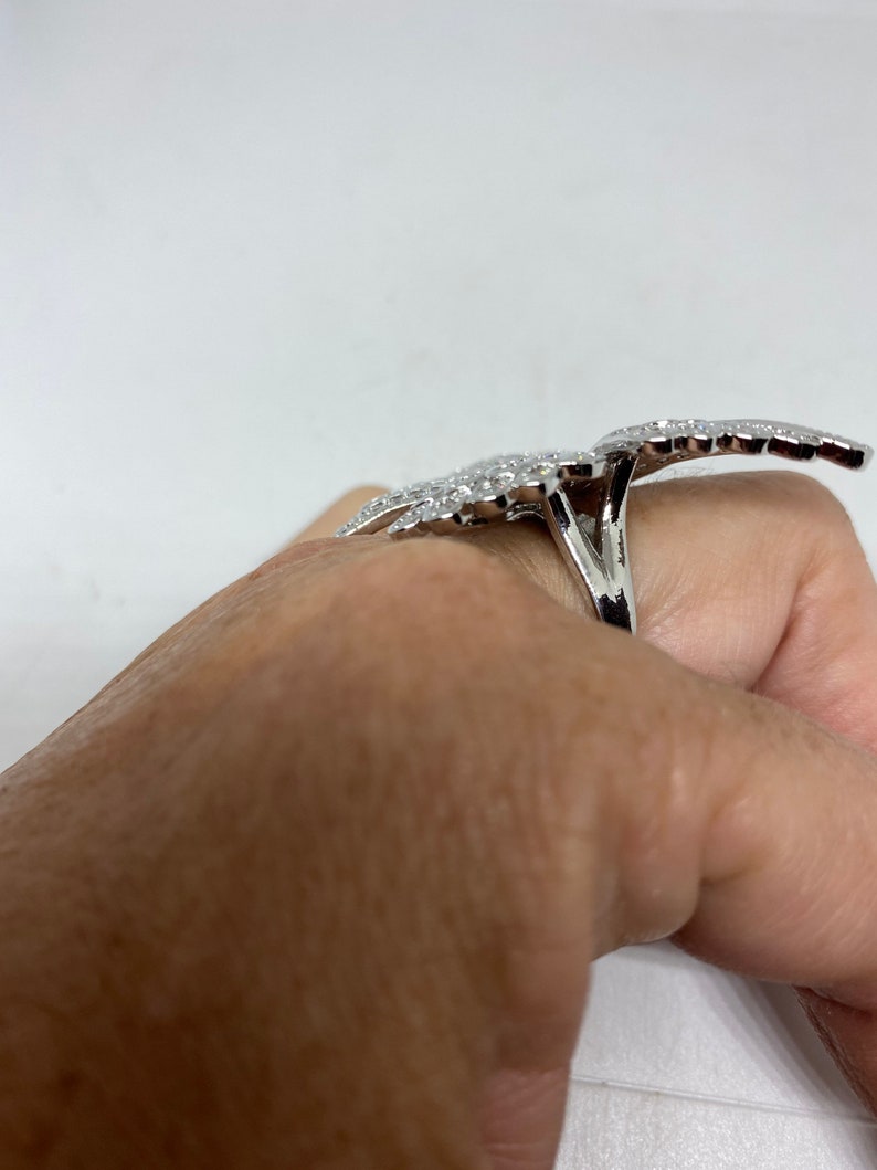 K&ouml;nnte beinhalten: Ein silberner Ring mit einem gro&szlig;en, komplizierten Design aus funkelnden Steinen. Der Ring wird an einem Finger getragen.