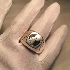 May include: A silver ring with a black and white design of a stylized animal head. The ring has a textured band with a wavy pattern.