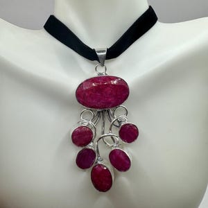 May include: A silver pendant necklace with a large oval ruby gemstone and six smaller ruby gemstones hanging from it. The necklace is on a black velvet choker.