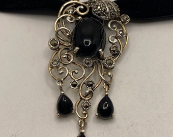Pode incluir: Um colar de choker de veludo preto com um pingente de prata que apresenta uma grande pedra preciosa preta cercada por filigranas intrincadas e acentos de marcasita. O pingente tem três gotas menores de pedras preciosas pretas penduradas nele.