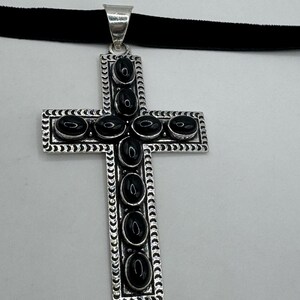 Op de afbeelding: Een zilveren kruis hanger met zwarte onyx stenen die in het midden en rond de randen zijn gezet. De hanger is bevestigd aan een zwarte fluwelen choker.