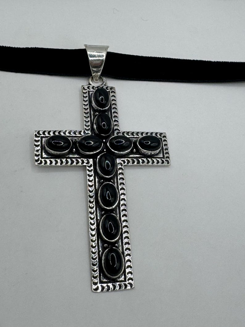 Op de afbeelding: Een zilveren kruis hanger met zwarte onyx stenen die in het midden en rond de randen zijn gezet. De hanger is bevestigd aan een zwarte fluwelen choker.