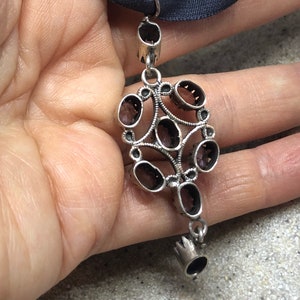 May include: A silver pendant with a cluster of oval-shaped, dark red gemstones set in a filigree design. The pendant hangs from a silver chain with a small, crown-shaped detail at the bottom.