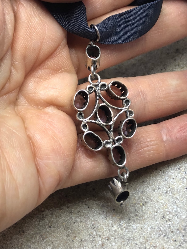 May include: A silver pendant with a cluster of oval-shaped, dark red gemstones set in a filigree design. The pendant hangs from a silver chain with a small, crown-shaped detail at the bottom.