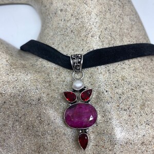 May include: A black velvet choker necklace with a silver pendant featuring a large oval pink gemstone surrounded by smaller red gemstones and a white pearl.