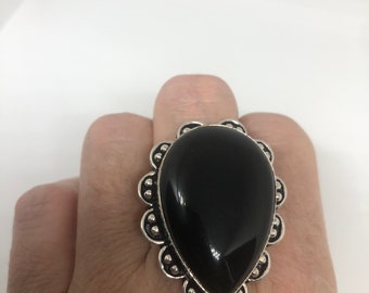May include: A large, teardrop-shaped black gemstone ring with a silver-colored, scalloped border. The ring's central stone is polished and reflects light. The setting has a detailed, ornate design. The ring is worn on a finger.