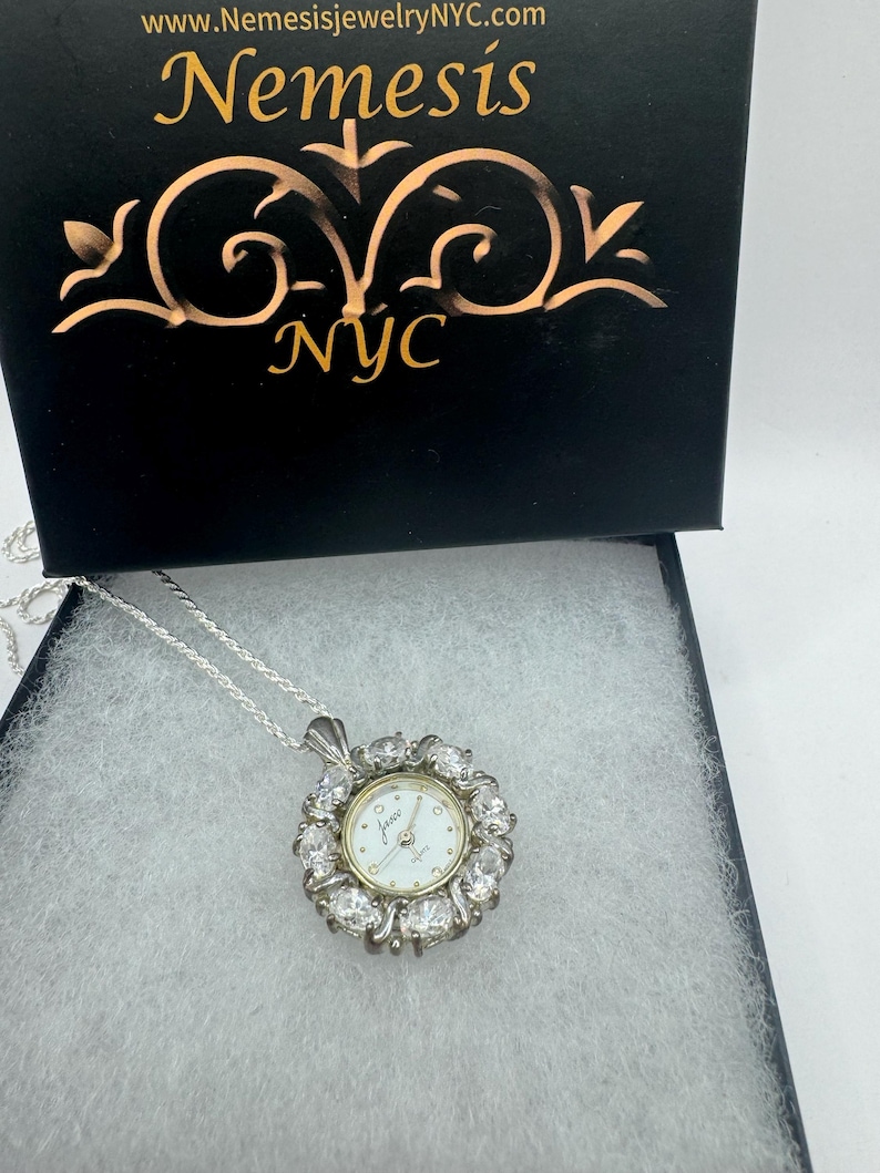 Op de afbeelding: Een zilveren ketting met een horlogehanger. De wijzerplaat is wit met goudkleurige wijzers en markeringen, omringd door heldere edelstenen. De hanger hangt aan een zilveren ketting, gepresenteerd in een zwarte doos met gouden letters "Nemesis NYC".