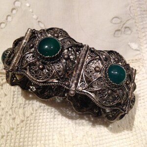 Puede incluir: Un brazalete de filigrana de plata con tres piedras verdes. El brazalete est&aacute; hecho de un delicado y complejo trabajo de metal y tiene un dise&ntilde;o de bisagra.