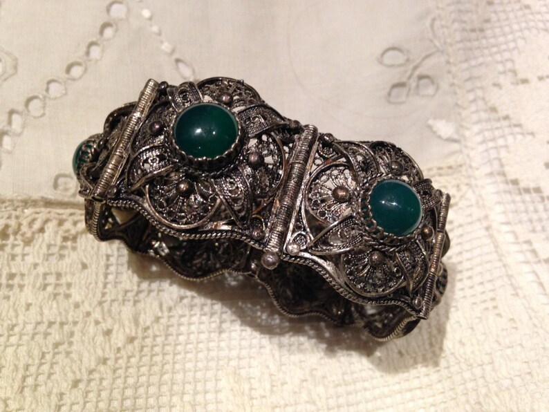 Puede incluir: Un brazalete de filigrana de plata con tres piedras verdes. El brazalete est&aacute; hecho de un delicado y complejo trabajo de metal y tiene un dise&ntilde;o de bisagra.