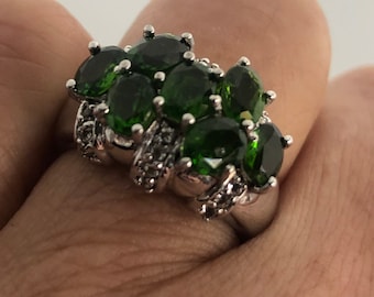 May include: A silver ring with a cluster of green gemstones and smaller clear stones. The ring is set on a finger.
