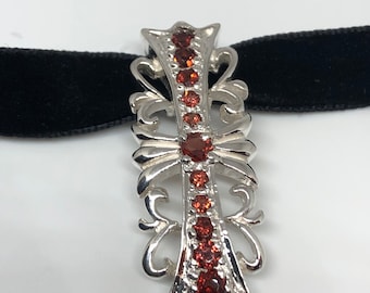 May include: A silver pendant with a cross design and red gemstones. The pendant is on a black velvet background.