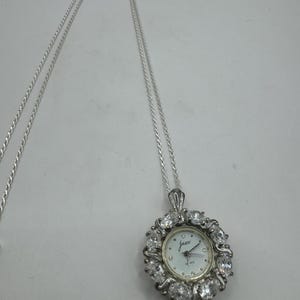 Op de afbeelding: Een zilverkleurige ketting met een hangerhorloge. De wijzerplaat van het horloge is wit met zwarte cijfers en wijzers, omringd door een halo van heldere kristallen. Het horloge draagt het merk "Jasco".