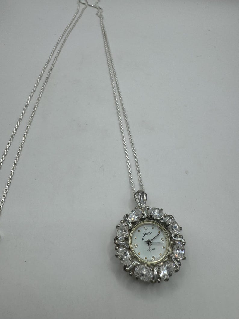 Op de afbeelding: Een zilverkleurige ketting met een hangerhorloge. De wijzerplaat van het horloge is wit met zwarte cijfers en wijzers, omringd door een halo van heldere kristallen. Het horloge draagt het merk "Jasco".